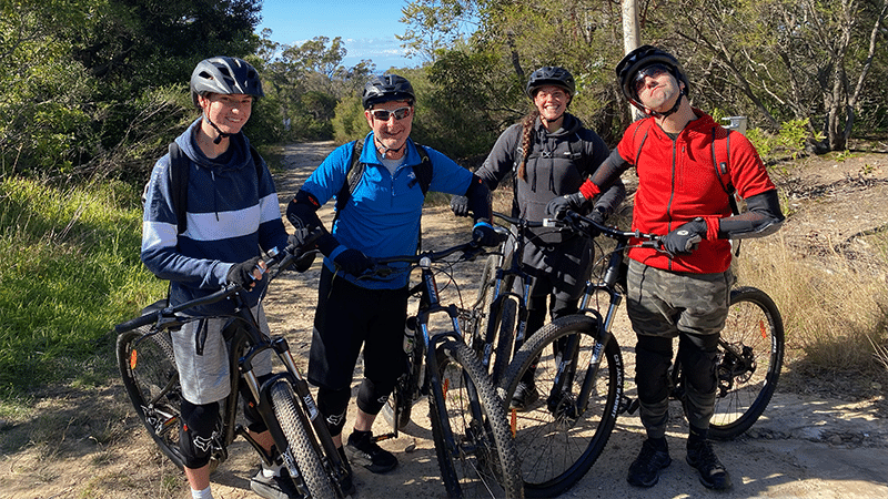 A group of 4 people in bike riding attire, standing together with their bikes in the bush